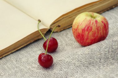 Two fresh cherries, apple and the open book on a linen fabric surface