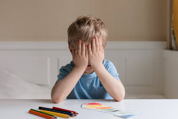 Милый маленький мальчик с светлыми волосами и голубой футболкой рисует цветные карандаши дома. Он положил руки на лицо и подумал о чем-то..