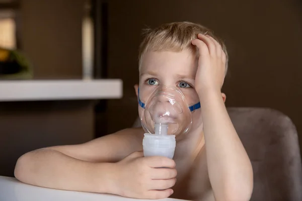 A sad little boy does inhalations with a nebulizer at home, he is sick. Help at home with cough, asthma. Concept health.