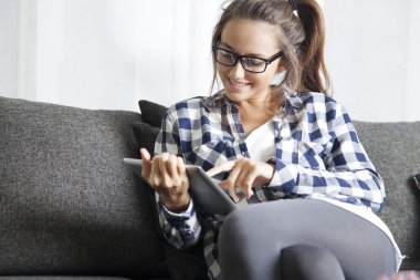 Oturma odasında kanepede oturan ve tablet kullanan genç bir kadın..