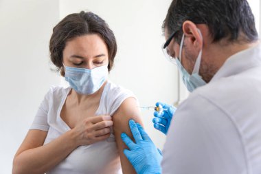Genç kadın ve doktoru maske takıyor ve bir doktorun ofisinde aşı oluyorlar.