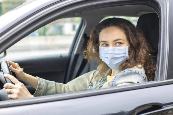 Young woman in a mask sitting in a car, protective mask against coronavirus, driver on a city street during a coronavirus outbreak.