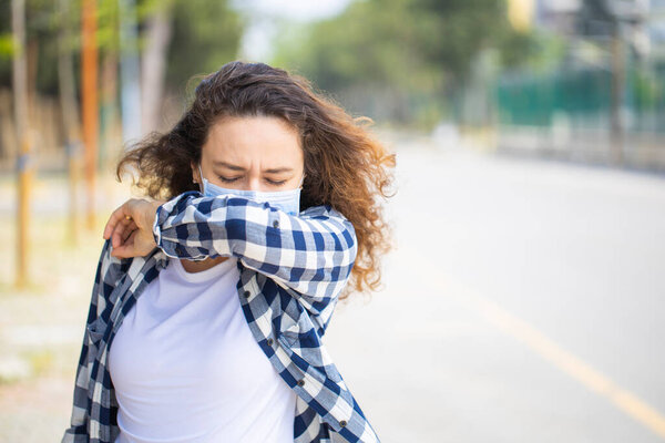 Woman with face mask sneezing into elbow while walking in the street