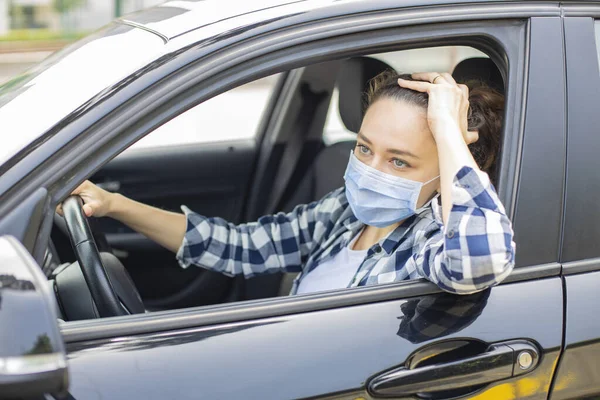 Young woman in a mask sitting in a car, protective mask against coronavirus, driver on a city street during a coronavirus outbreak.