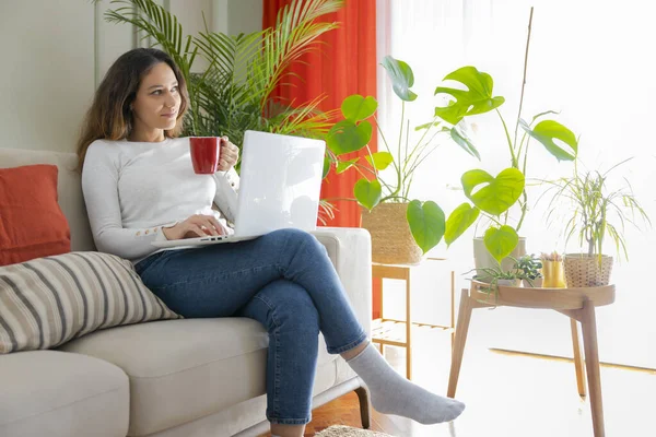 年轻妇女坐在沙发上使用笔记本电脑 晴天在客厅喝咖啡 — 图库照片