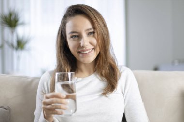 Evdeki kanepede su içen genç bir kadın. Yeterince su içmenin sağlık yararları. 