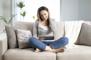 Oturma odasında kanepede oturan ve tablet kullanan genç bir kadın..