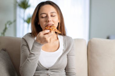 Genç bir kadın evde bir dilim pizza yiyor. Çekici genç bir kadın oturma odasında lezzetli pizzalar yiyor..