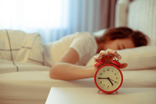 Wake up of an asleep young woman stopping red alarm clock on the bed in the morning