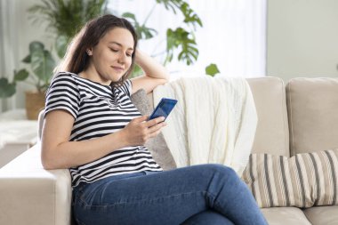 Evde koltukta otururken cep telefonu kullanan genç bir kadın. 