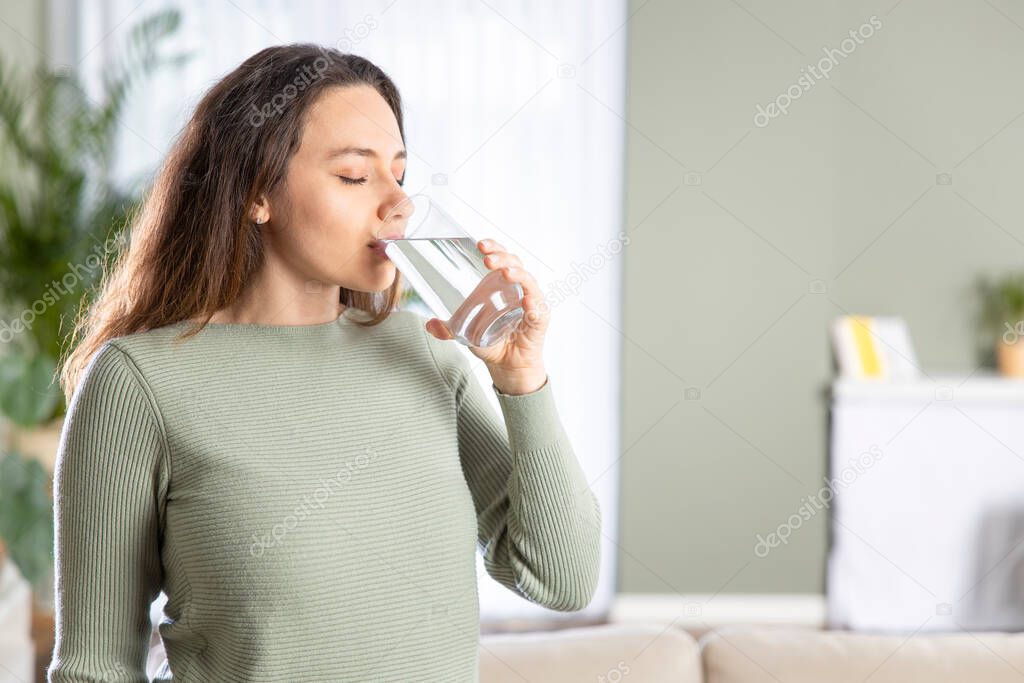 Young woman drinking water sitting on a couch at home. Health benefits of drinking enough water concepts. 