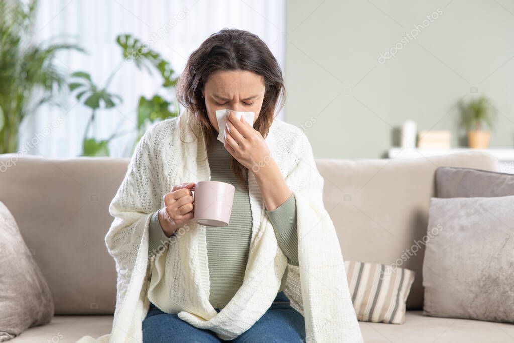 Sick young woman blowing the nose using tissue paper while sitting on the sofa at home