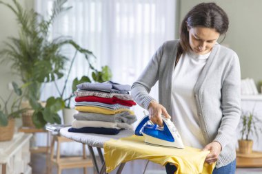 Genç bir kadın oturma odasındaki kıyafetleri ütülüyor. Katlanmış giysiler ütü masasında duruyor. 