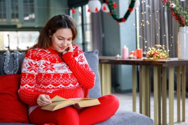 Kırmızı şenlikli süveter giyen genç bir kadın kanepeye oturup kitap okuyor. Noel süslemeleri, rahat bir tatil evi ve rahatlama sahnesi