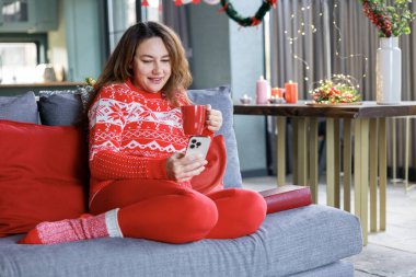 Neşeli kadın gri bir koltukta oturuyor, kırmızı tatil süveteri giyiyor, telefonunu kontrol ederken kırmızı bardaktan içiyor. Şenlikli dekor ve sıcak ışıklandırma. Ev yapımı Noel havası