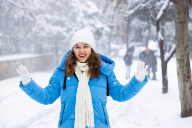 Mavi ceketli beyaz örgü şapkalı ve atkılı genç kadın taze karda duruyor, kolları geniş, karlar etrafını sararken gülümsüyor.