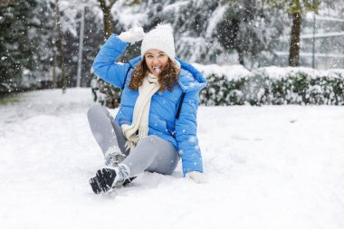 Mavi kışlık ceketli, karlı bir kış gününde kartoplarıyla oynayan neşeli genç bir kadın. Huzurlu bir parkın etrafına yavaşça kar yağar