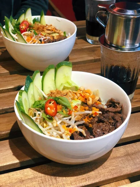 Vietnamese beef noodle bowl (Bun Bo Xao) with fresh cucumber, chili slice & soy bean sprouts Side view two plates with traditional asian food on wooden table. Delicious authentic food in Vietnam.