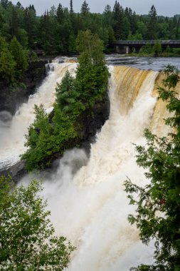 Güçlü şelaleler, yemyeşil ağaçlar ve uzaktaki bir köprüyle çevrili koyu kayaların üzerine dökülüyor..