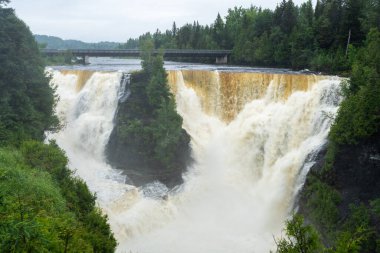 Güçlü bir şelale bir uçurumdan aşağı dökülüyor, üzerinde nehri kaplayan bir köprü var..