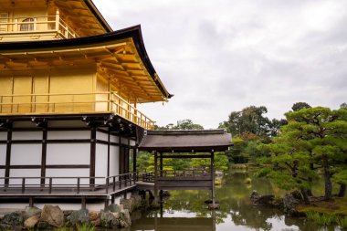 Altın bir pavyon, yemyeşil yemyeşil ve bulutlu gökyüzü ile çevrili sakin bir göletin yanında oturur..
