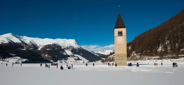 Steeple Curon Venosta och sjön Resia i vinter med snö, med människor, blå himmel med moln, Dolomiterna, Italien — Stockfoto