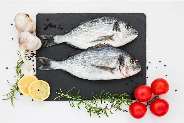 Poisson dorado frais sur planche à découper en ardoise, romarin, ail, citron et tomates. Vue du dessus — Photo