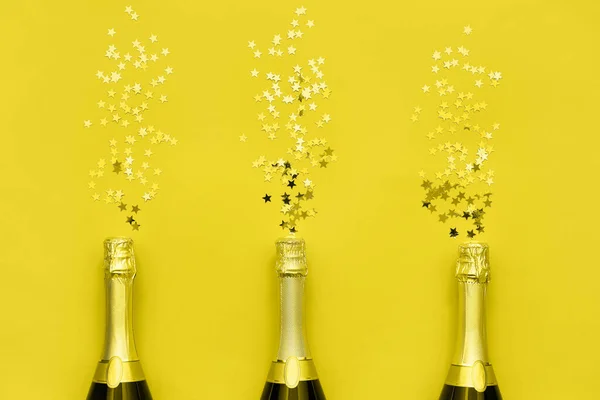 Tres botellas de champán con estrellas de confeti sobre fondo amarillo. Copiar espacio, vista superior — Foto de Stock