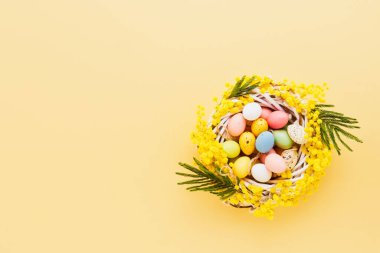 Sarı arka planda renkli Paskalya yumurtaları ve mimoza çelengi. Üst görünüm, alanı kopyala