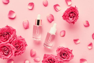 Tıbbi ve kozmetik kullanım için iki şeffaf cam damlası şişesi ve pembe arka planda pembe güller. SPA konsepti. Üst görünüm, metin için boşluk kopyala