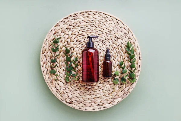 Dropper bottle of essential oil or serum, eucalyptus and dark dispenser bottle of shower gel, on rattan background. Beauty and SPA concept. Flat lay, copy space, toned