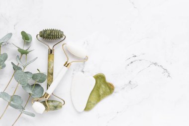 Masaj aletleri. Guasha masaj aleti, yeşim yüz silindirleri ve beyaz mermer arka planda okaliptüs dalları. SPA konsepti.