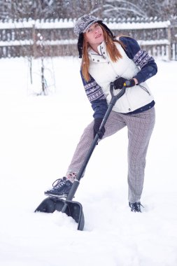 Yüzünde bir gülümseme olan güzel bir kız evin bahçesindeki karı temizliyor. Kürekle kar temizleme.