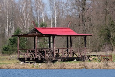 Kırmızı çatılı ahşap bir çardak ormanın yanında duruyor..