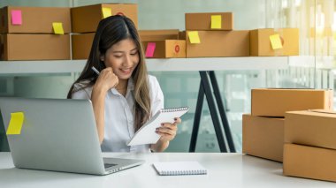 Bağımsız genç Asyalı kadınlar evde çalışıyor ve KOBİ girişimcilerine İnternet üzerinden başlatıyorlar. Sipariş sayısını kontrol etmek ve not defterine yazmak için dizüstü bilgisayar kullananlar.