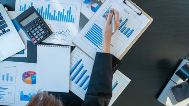 Bir iş kadınının eli masanın üstünde dizüstü bilgisayarı ve hesap makinesi olan bir grafiği işaret ediyor.