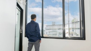 Başarılı, uzun mavi gömlekli genç bir işadamı bir ofis binasının en üst katında duruyor ve büyük pencerelerden dışarı bakıyor..