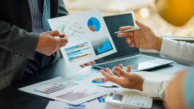 İki işadamı, konferans odasında hesap makinesi ve turuncu ışıklı bir dizüstü bilgisayarla bir masanın üzerindeki maliyet grafiğini inceliyorlardı..