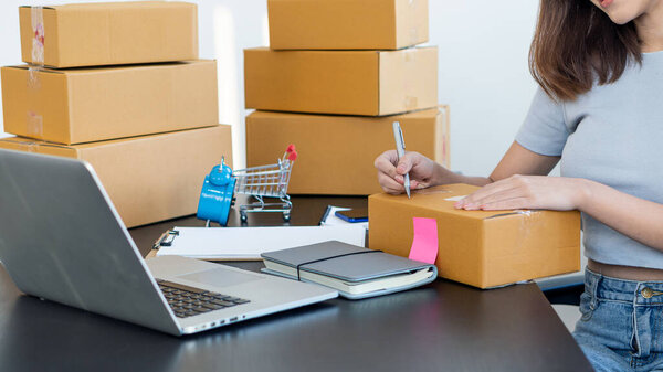 Young Asian businesswoman starts a small business, an online SME owner using a laptop, customizes customer orders with packaging boxes at home office and delivers their products online.