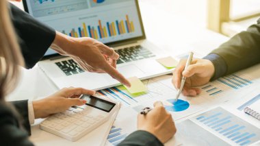 İş ekibi toplantı odasındaki dizüstü bilgisayarlarla masalarının üzerindeki maliyet grafiklerini analiz ediyor..