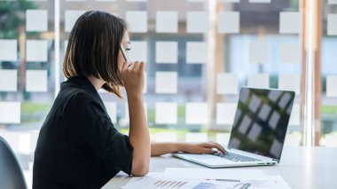 Genç Asyalı iş kadını, dokümanlar, grafikler ve dizüstü bilgisayarlar üzerinde çalışırken ofiste gülümsüyor..