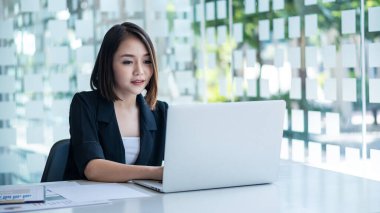 Çekici bir Asyalı iş kadını masa başında oturuyor ve ofiste dokümanlar, grafikler ve dizüstü bilgisayarlarla çalışıyor..