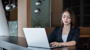 Çekici bir Asyalı iş kadını masa başında oturuyor ve ofiste dokümanlar, grafikler ve dizüstü bilgisayarlarla çalışıyor..