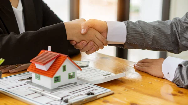 A young businessman shakes hands with a partner after Zen, a real estate investment contract document on a table with a house design and documents and money and a laptop.