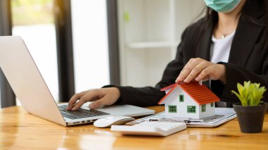 Bir emlakçı müşterilerine kontrat belgeleri, dizüstü bilgisayar ve dolar banknotları olan bir ev düzeni sunar. Ev satın almakla ilgilenen müşteriler ev satın alma sözleşmesini tamamlamak için imzalarını atarlar..