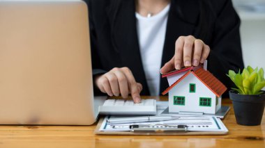 Bir emlakçı müşterilerine kontrat belgeleri, dizüstü bilgisayar ve dolar banknotları olan bir ev düzeni sunar. Ev satın almakla ilgilenen müşteriler ev satın alma sözleşmesini tamamlamak için imzalarını atarlar..