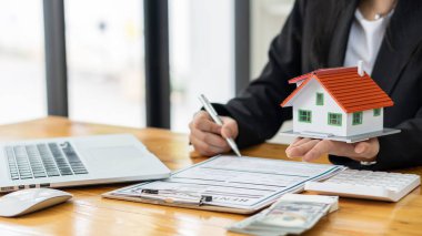 Bir emlakçı müşterilerine kontrat belgeleri, dizüstü bilgisayar ve dolar banknotları olan bir ev düzeni sunar. Ev satın almakla ilgilenen müşteriler ev satın alma sözleşmesini tamamlamak için imzalarını atarlar..