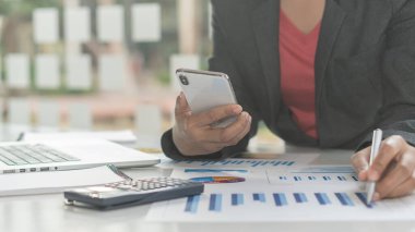 Masada oturan ve akıllı telefon kullanan bir iş kadını masanın üstünde bir dizüstü bilgisayar ve ofis çizelgeleri ve grafikleri var..