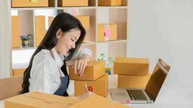 Asyalı kız başlangıç noktası olarak kameraya bakıyor, ev ve dizüstü bilgisayarla çalışıyor KOBİ 'ler, tedarik zincirleri, tedarik, dış ticaret, omnichannel, çevrimiçi satış kavramları gibi paketleri hazırlıyor..