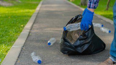Genç bir adam, çevre için bahçeleri temizlemek için çöp torbasında plastik şişe topluyor..
